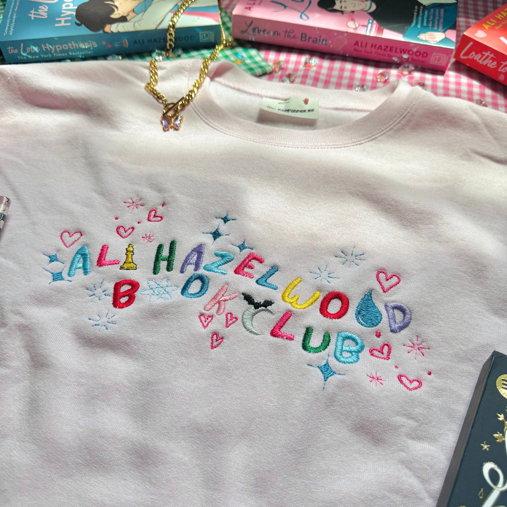 Pale Pink sweatshirt with colorful text 'Ali Hazelwood Book Club' on a surface with books in the background.