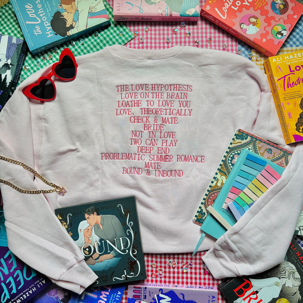Pale Pink sweatshirt with Ali Hazelwood's bibliography embroidered, surrounded by books and a notebook on a checkered surface.