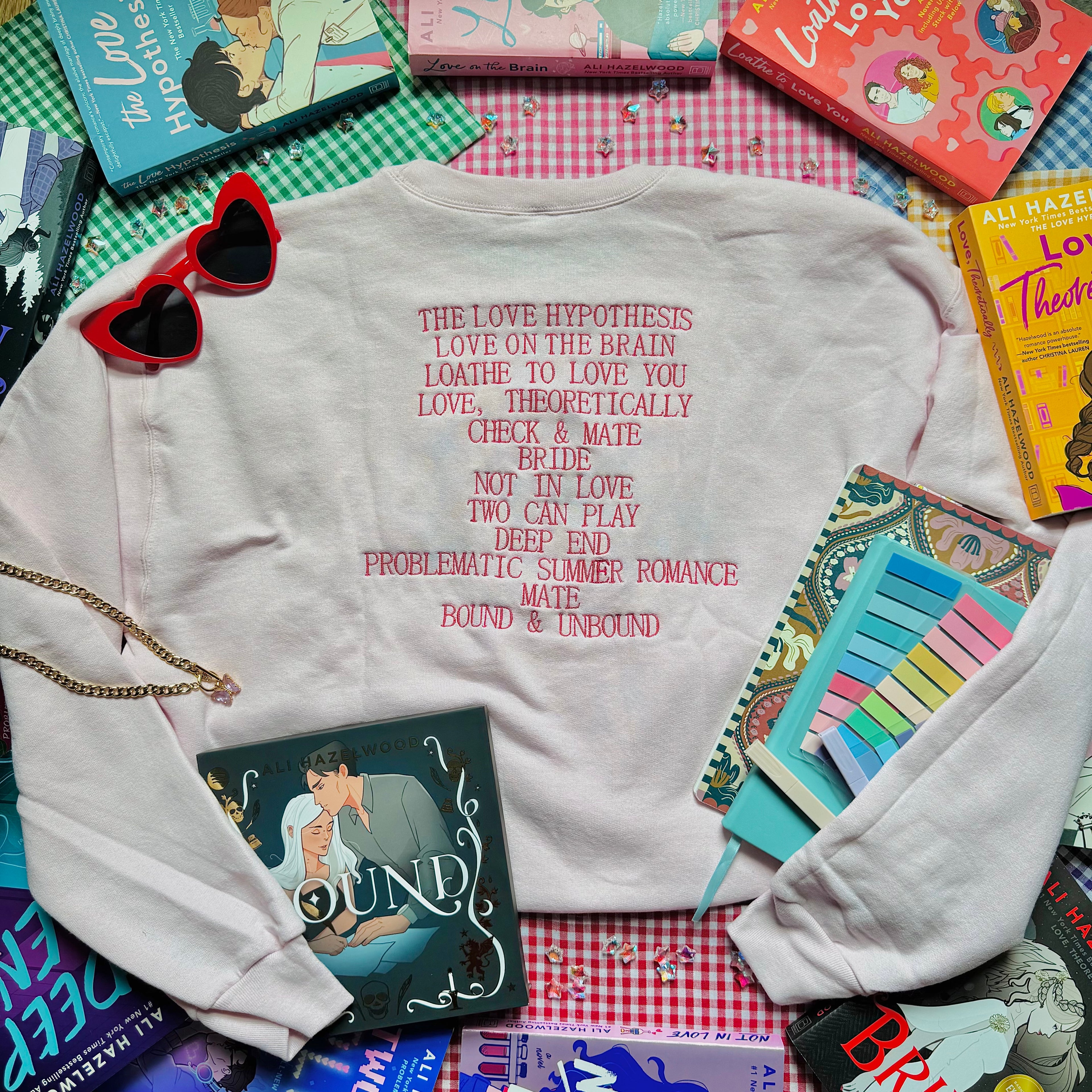 Pale Pink sweatshirt with Ali Hazelwood's bibliography embroidered, surrounded by books and a notebook on a checkered surface.