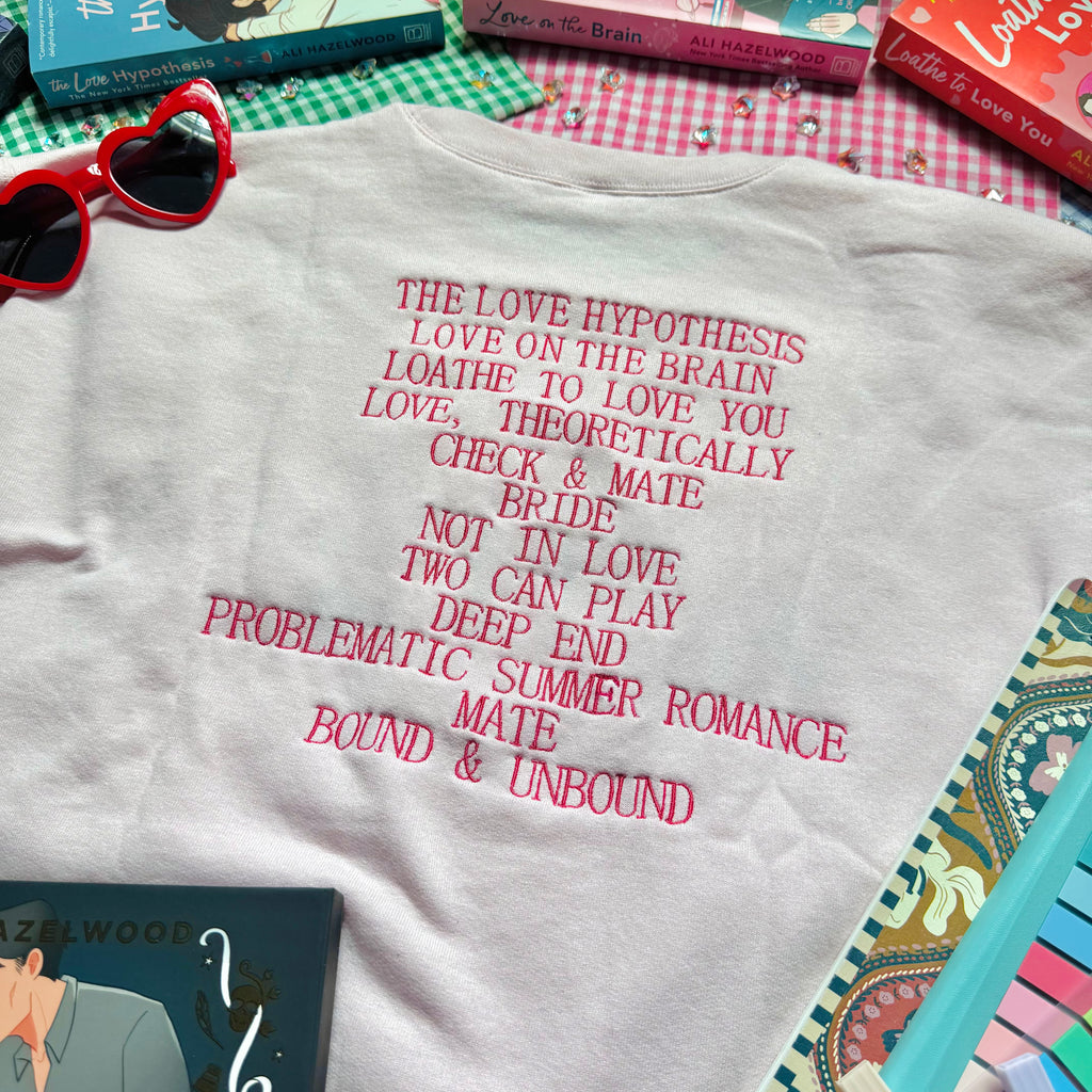 Pale Pink sweatshirt with Ali Hazelwood's bibliography embroidered, surrounded by books and a notebook on a checkered surface.