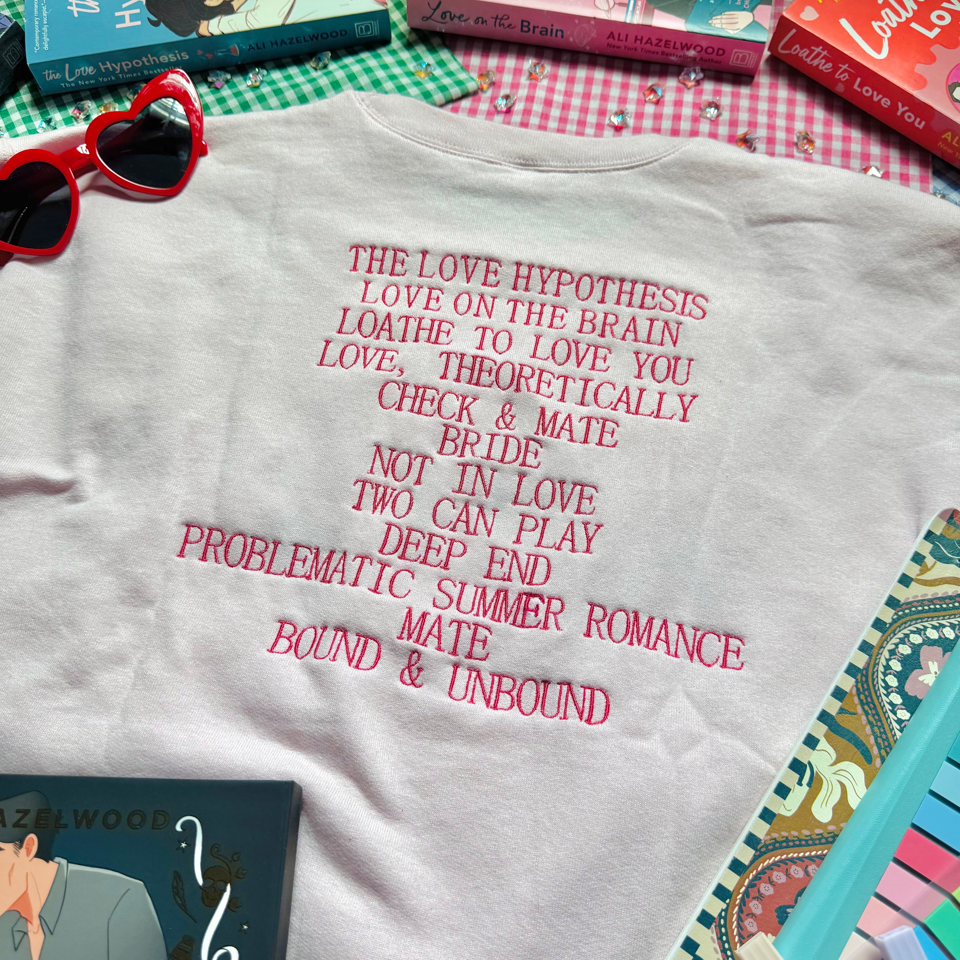 Pale Pink sweatshirt with Ali Hazelwood's bibliography embroidered, surrounded by books and a notebook on a checkered surface.