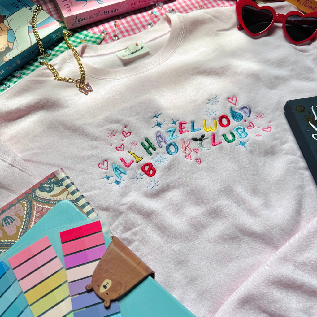 Pale Pink sweatshirt with 'Ali Hazelwood Book Club' text, surrounded by books and a notebook on a checkered surface.