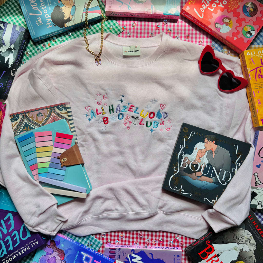 Pale Pink sweatshirt with 'Ali Hazelwood Book Club' text, surrounded by books and a notebook on a checkered surface.