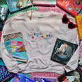 Pale Pink sweatshirt with 'Ali Hazelwood Book Club' text, surrounded by books and a notebook on a checkered surface.