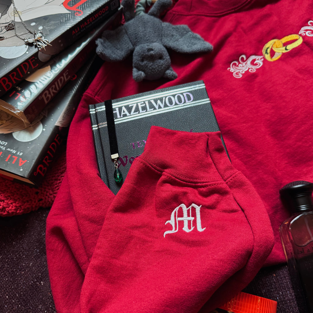 Red sweatshirt with a monogram, books, and a small plush toy on a dark surface