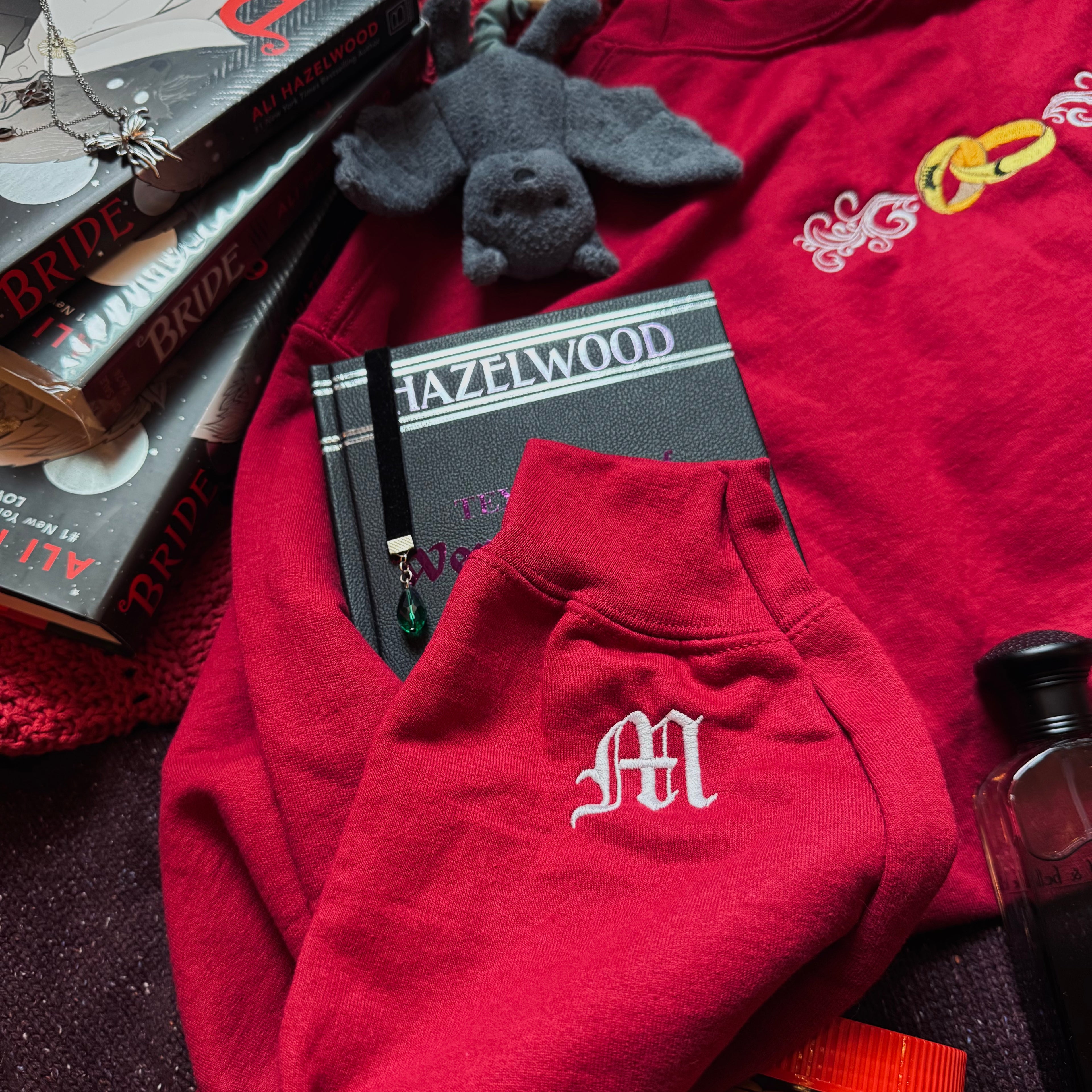 Red sweatshirt with a monogram, books, and a small plush toy on a dark surface