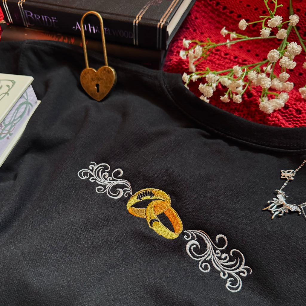 Black fabric with gold rings and white flowers embroidery, books, and a heart-shaped keychain in the background.