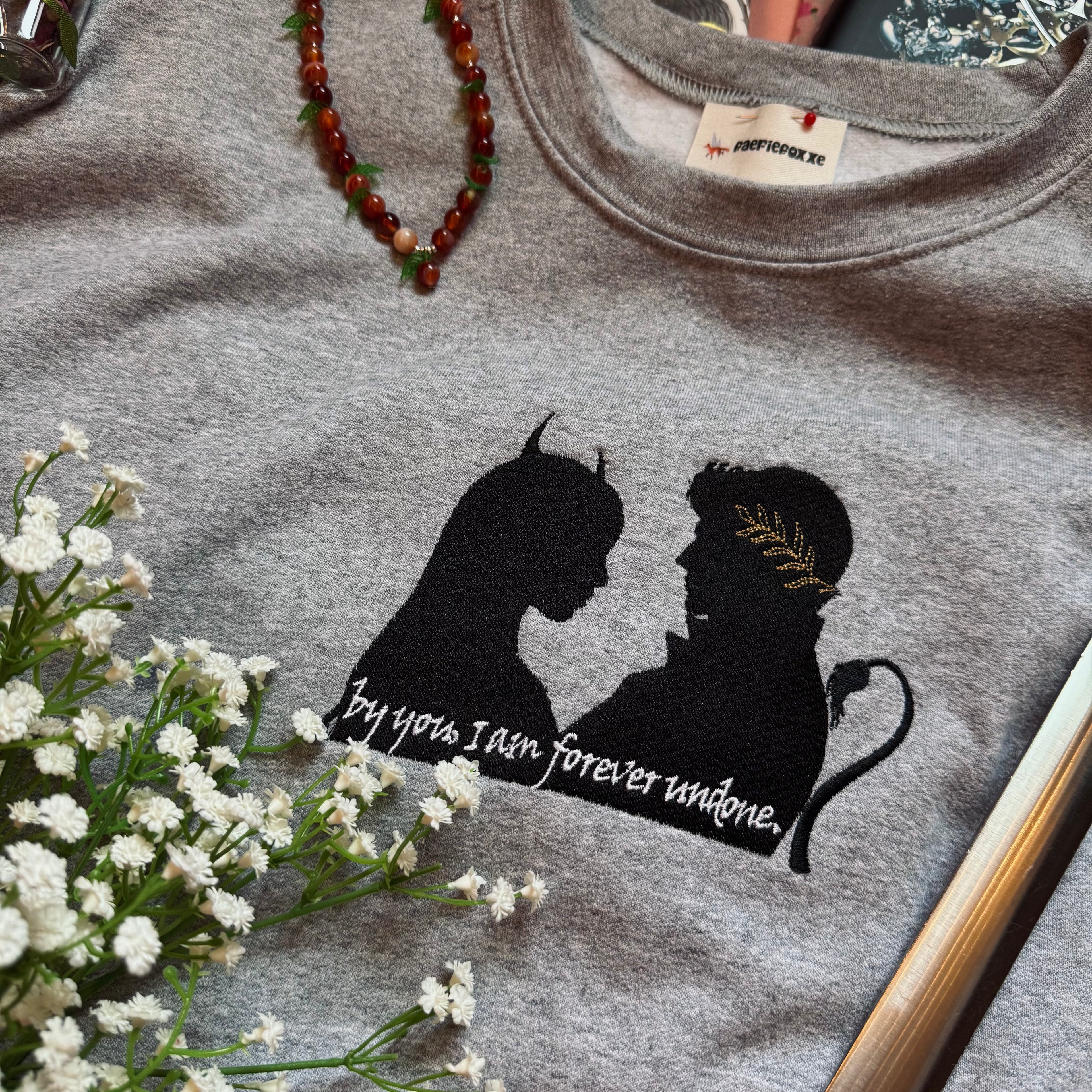 Gray sweatshirt with a silhouette design and text, surrounded by flowers and a necklace.
