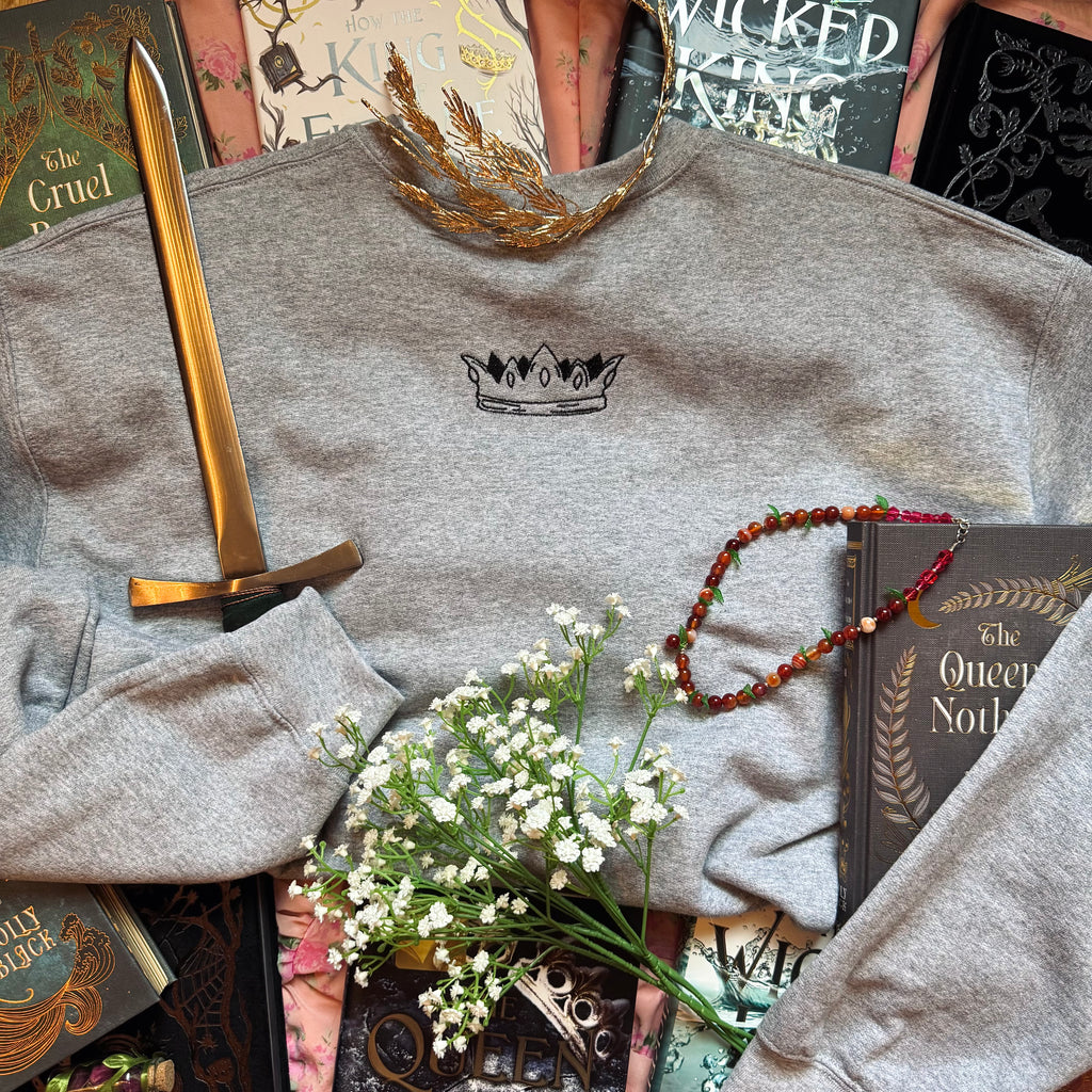 Gray sweatshirt with a crown logo, gold sword, tiara, necklace, and books on a textured surface.
