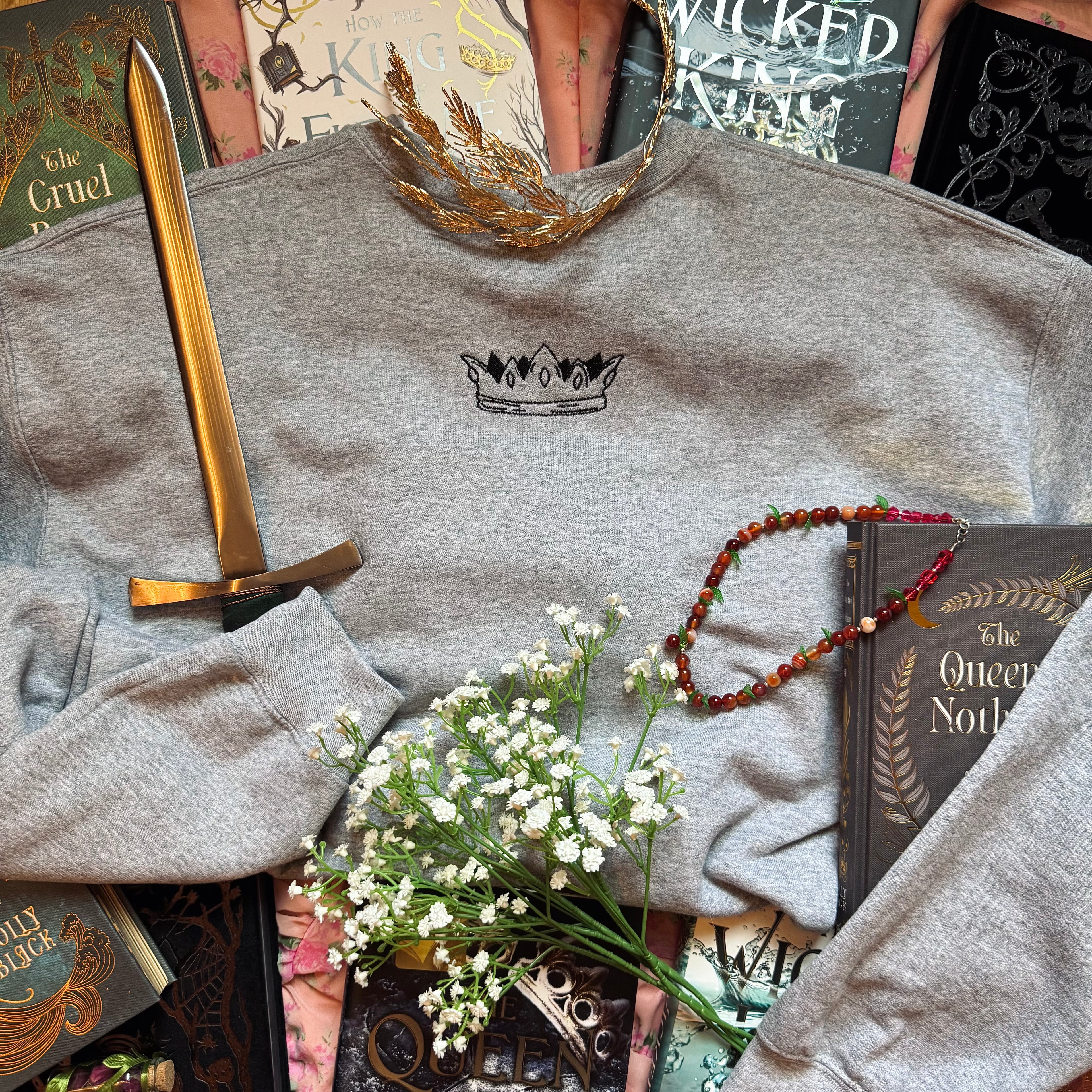 Gray sweatshirt with a crown logo, gold sword, tiara, necklace, and books on a textured surface.