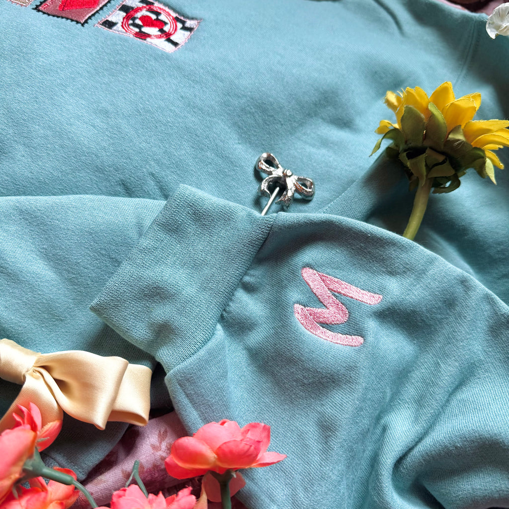 Teal sweatshirt with embroidered "M" design on sleeve.