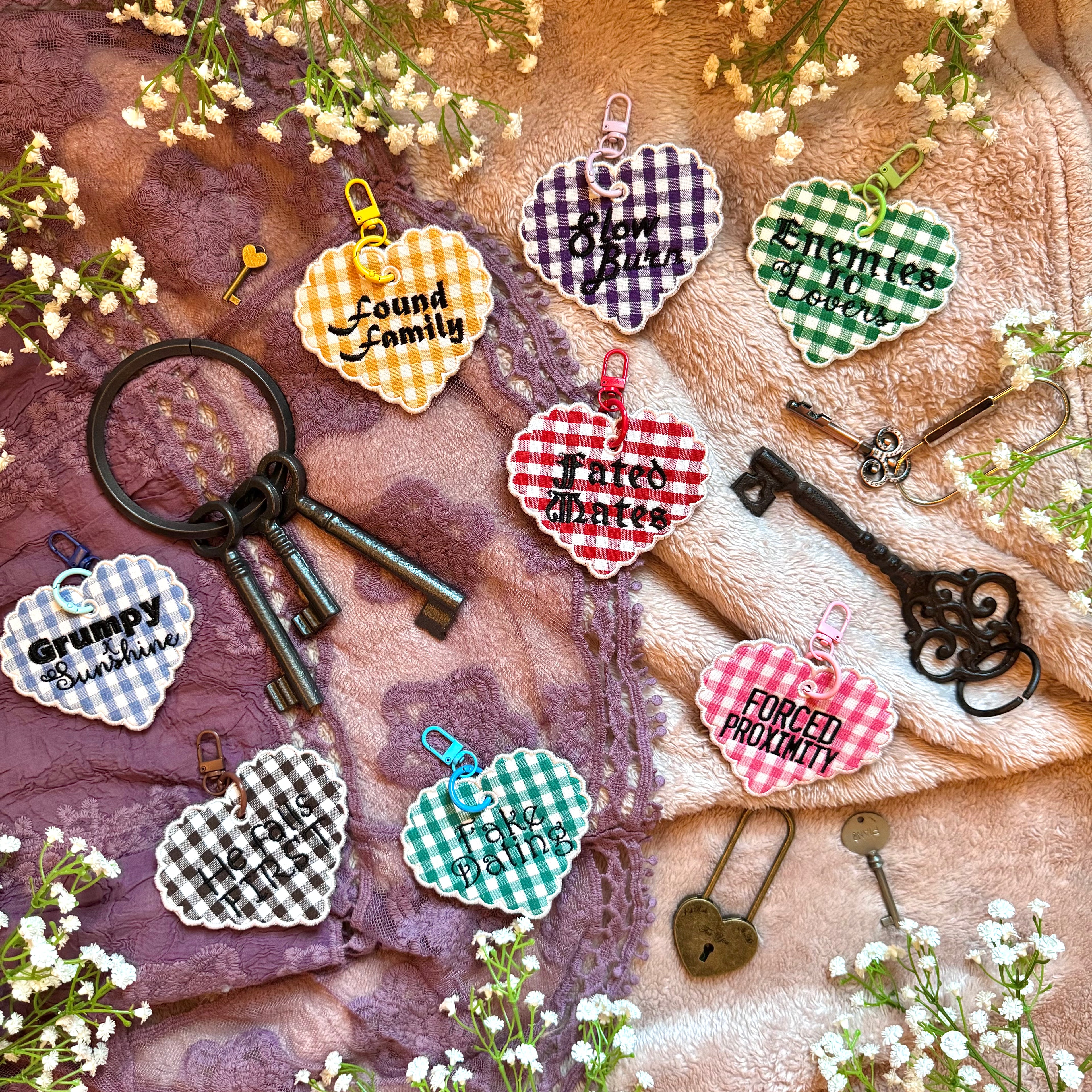 Heart-shaped keychains with romance tropes text on a textured surface with keys and flowers.