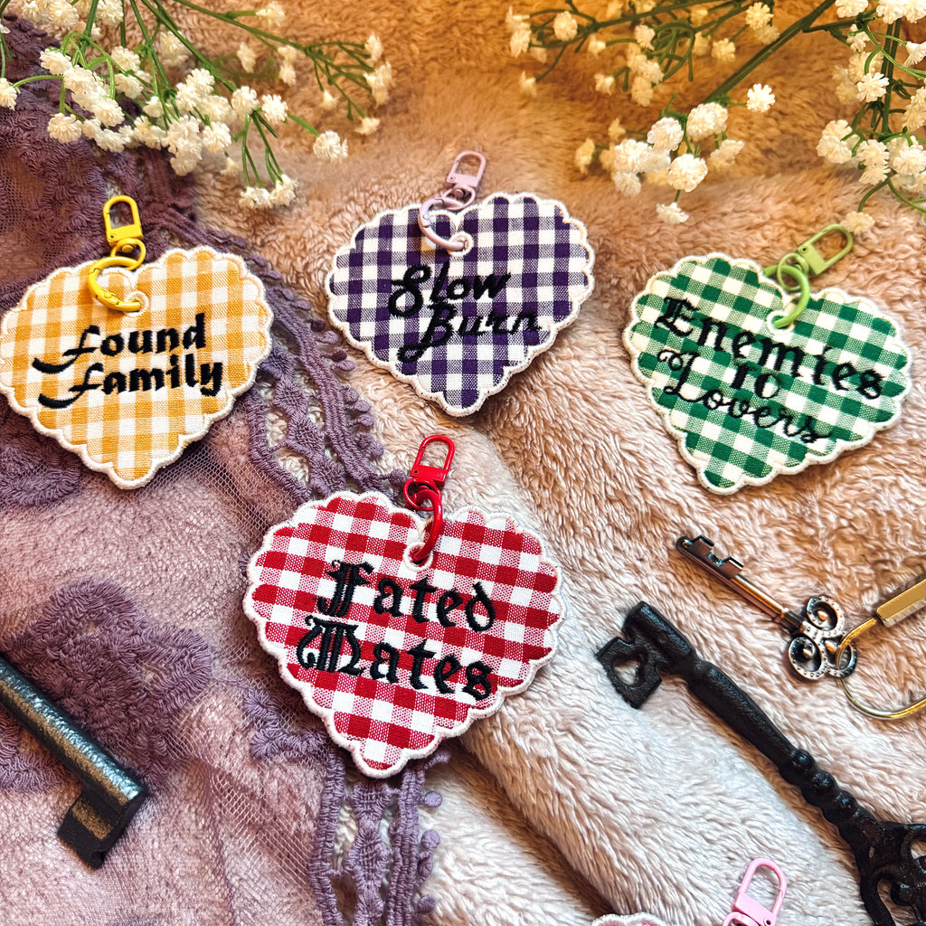 Decorative heart-shaped keychain with romance trope text, surrounded by vintage keys and flowers on a textured fabric background.