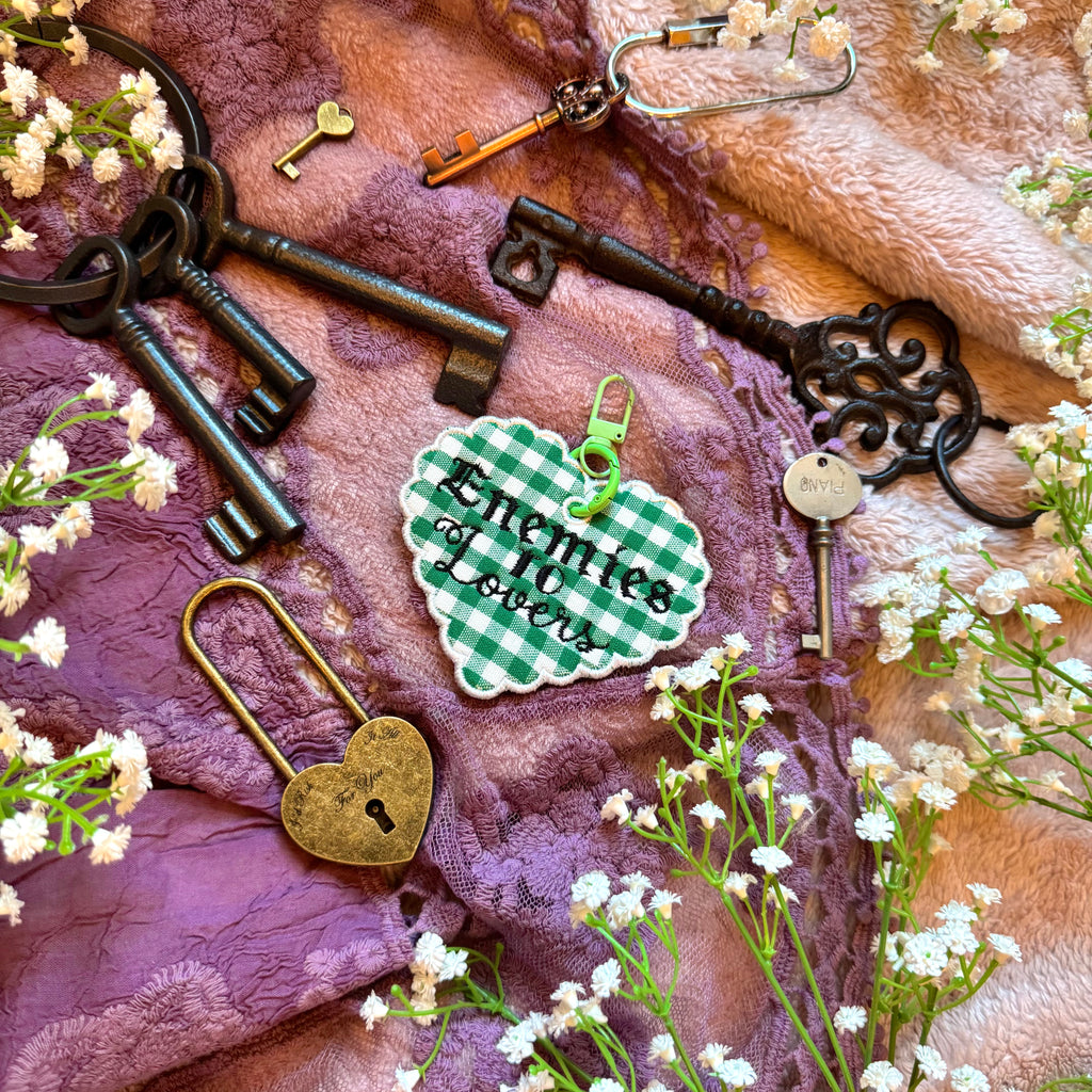 Decorative heart-shaped keychain with 'Enemies to Lovers' text, surrounded by vintage keys and flowers on a textured fabric background.
