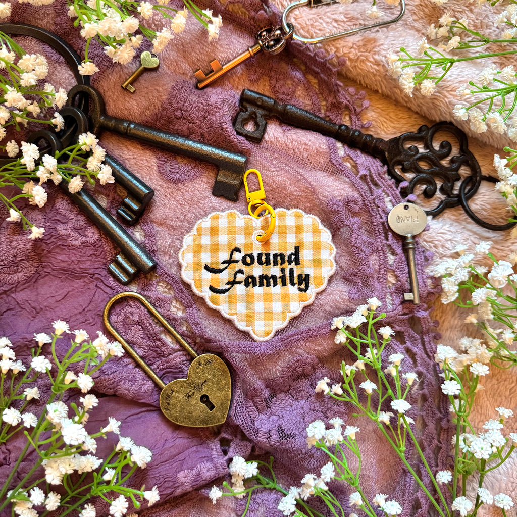 Decorative heart-shaped keychain with 'Found Family' text, surrounded by vintage keys and flowers on a textured fabric background.