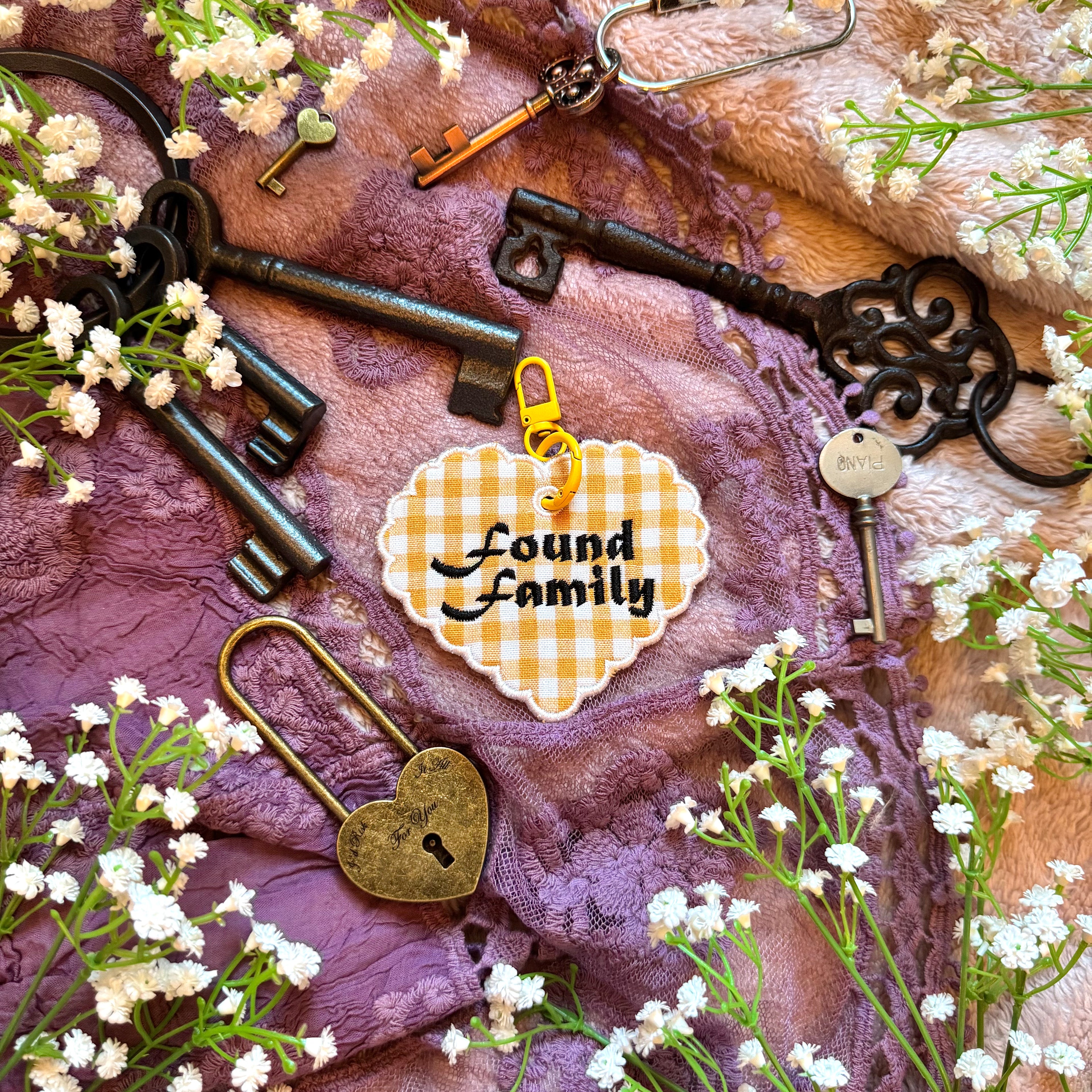 Decorative heart-shaped keychain with 'Found Family' text, surrounded by vintage keys and flowers on a textured fabric background.