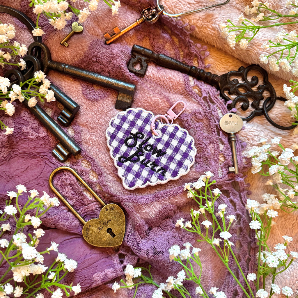 Decorative heart-shaped keychain with 'Slow Burn' text, surrounded by vintage keys and flowers on a textured fabric background.