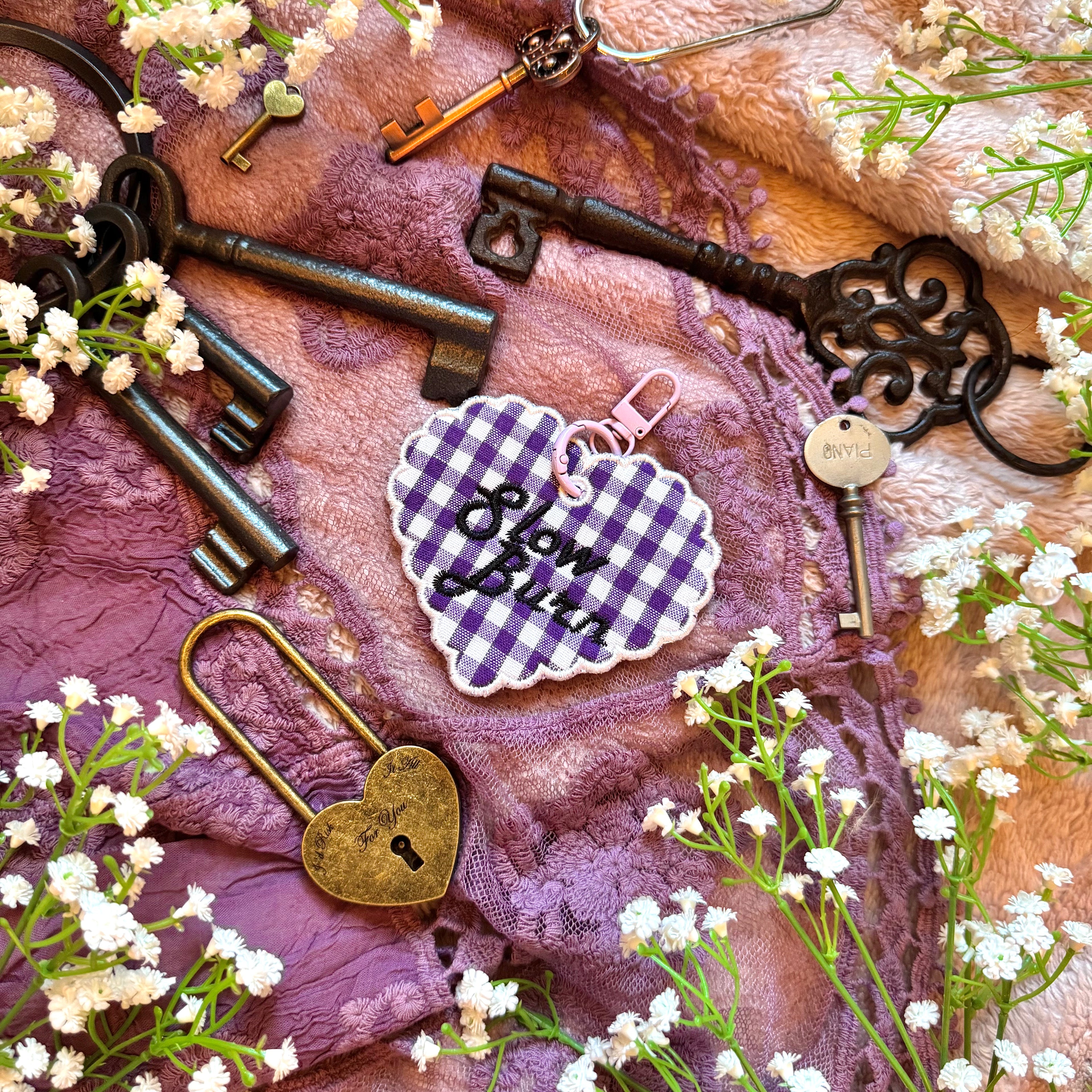 Decorative heart-shaped keychain with 'Slow Burn' text, surrounded by vintage keys and flowers on a textured fabric background.