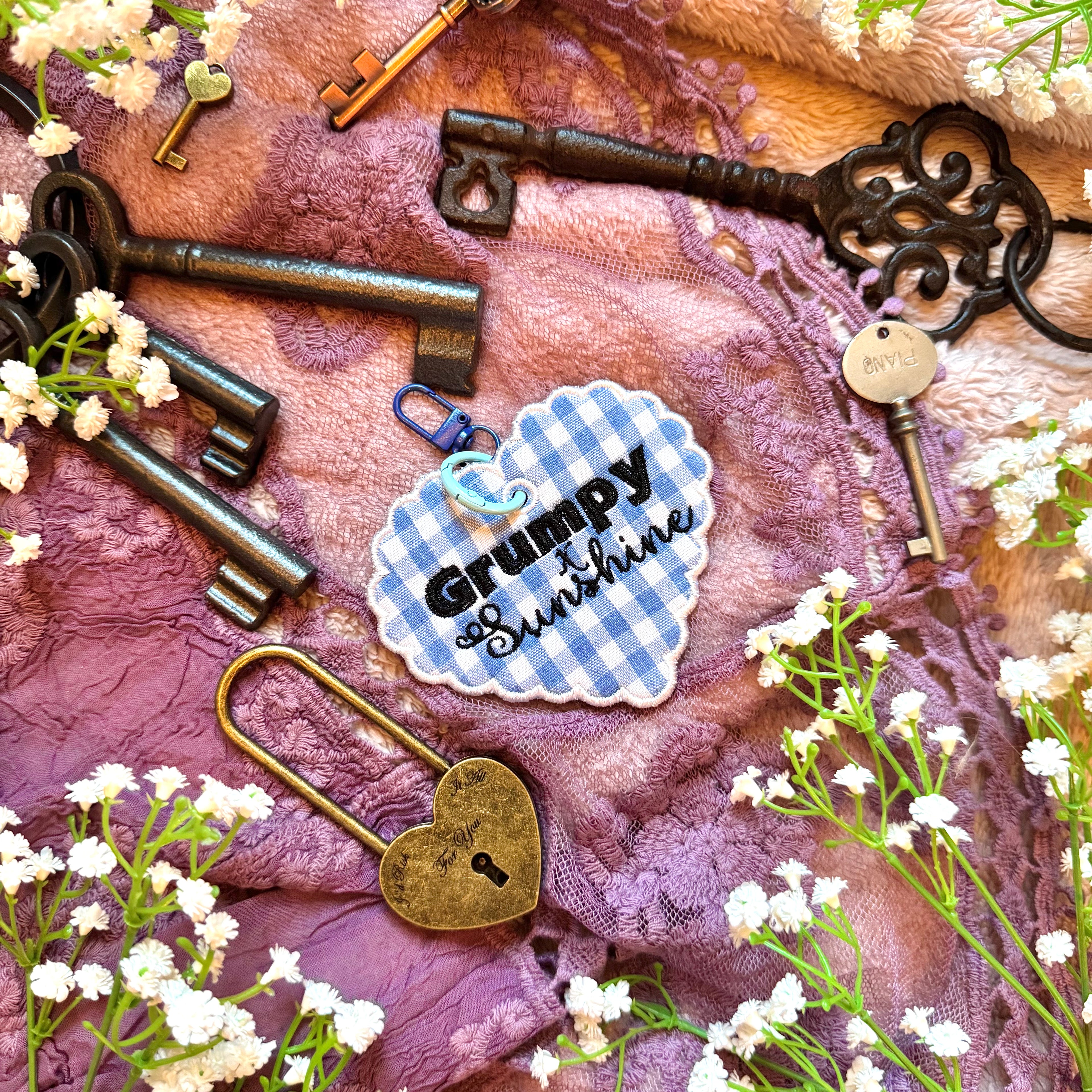 Decorative heart-shaped keychain with 'Grumpy x Sunshine' text, surrounded by vintage keys and flowers on a textured fabric background.
