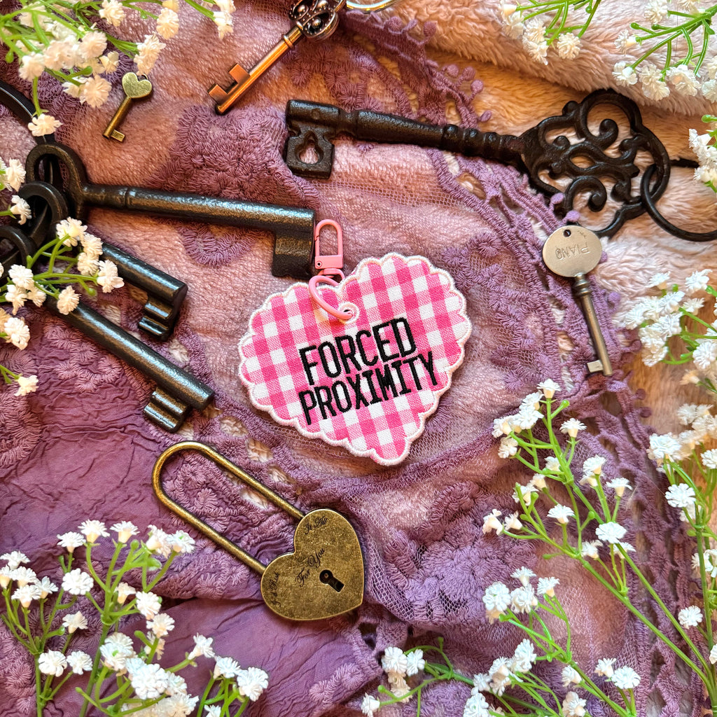 Decorative heart-shaped keychain with 'Forced Proximity' text, surrounded by vintage keys and flowers on a textured fabric background.