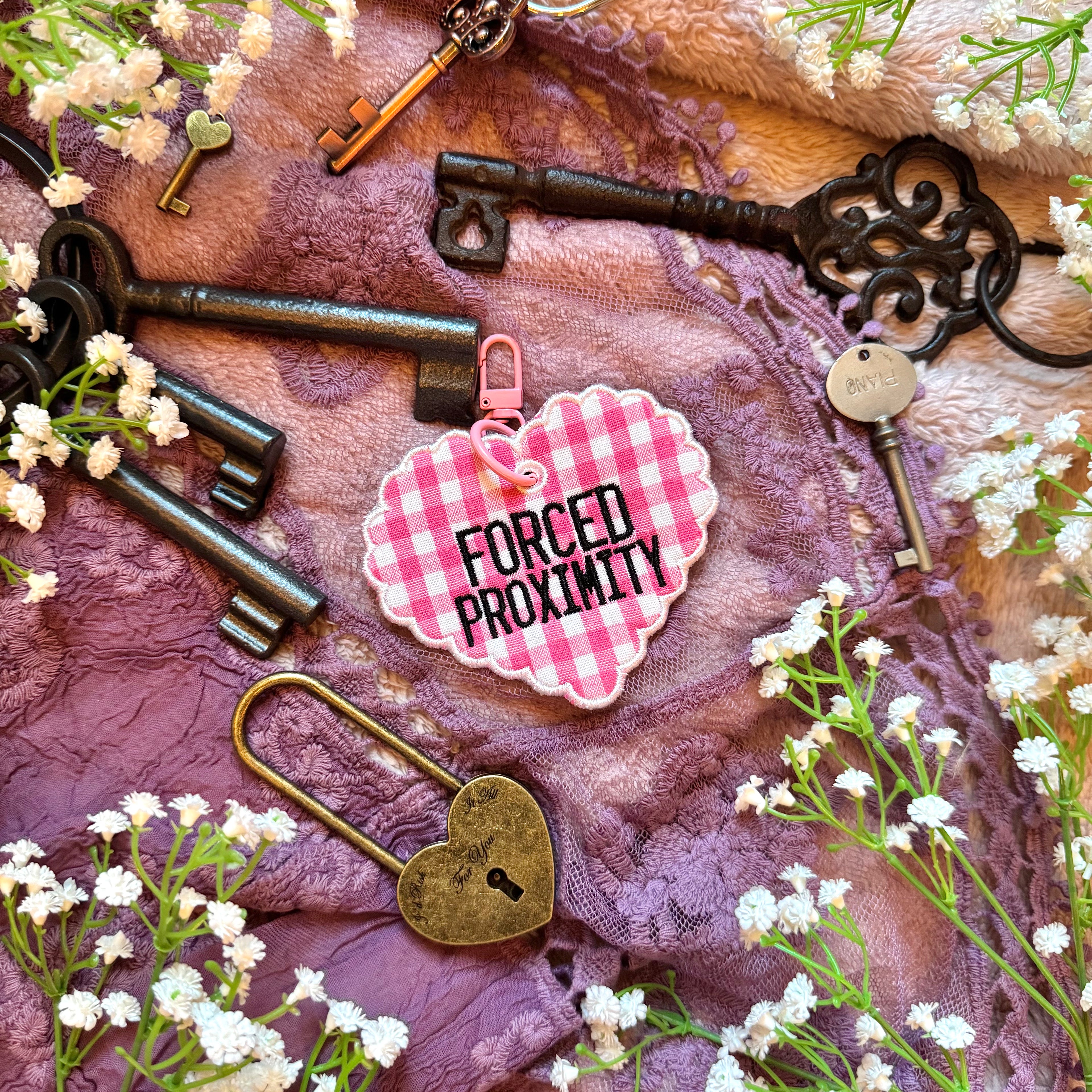 Decorative heart-shaped keychain with 'Forced Proximity' text, surrounded by vintage keys and flowers on a textured fabric background.