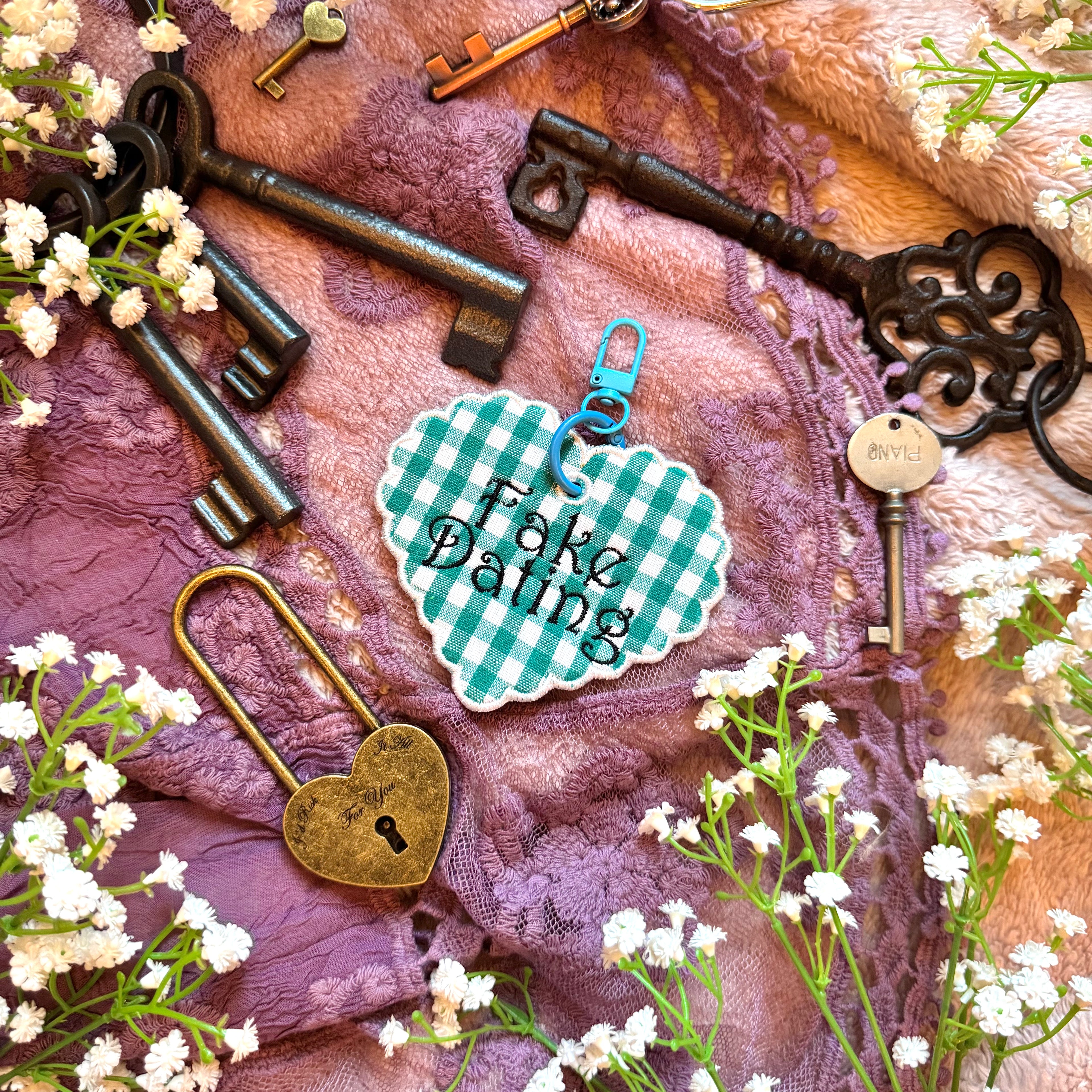 Decorative heart-shaped keychain with 'Fake Dating' text, surrounded by vintage keys and flowers on a textured fabric background.