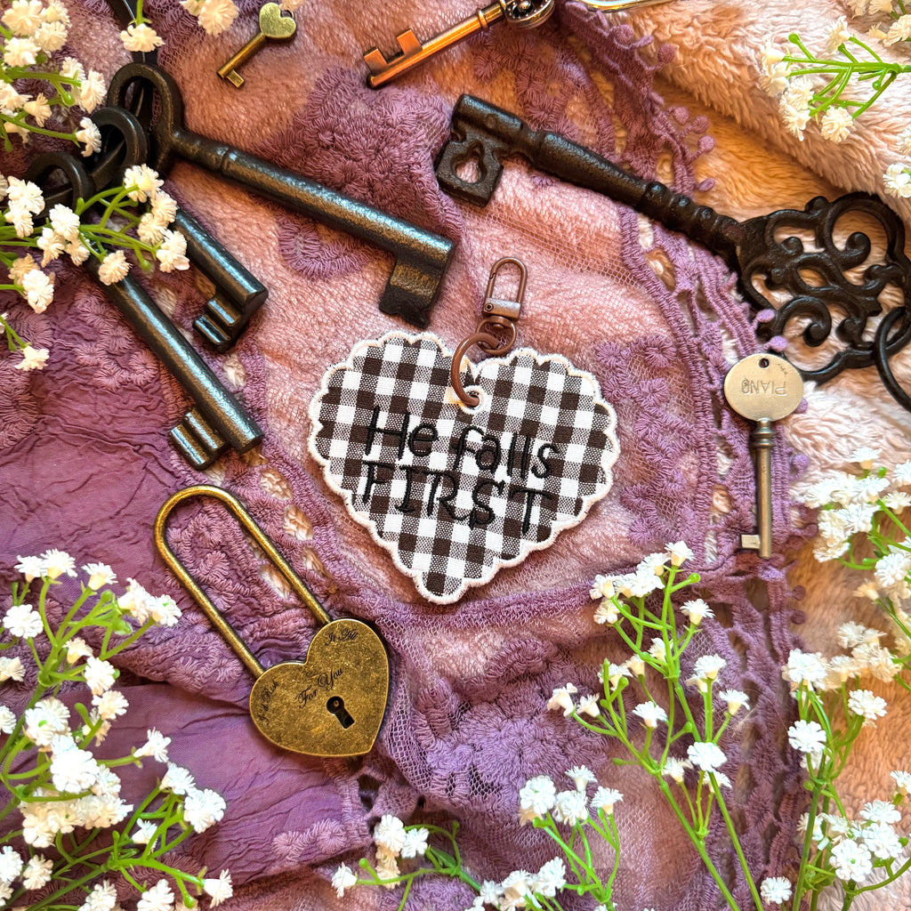 Decorative heart-shaped keychain with 'He Falls First' text, surrounded by vintage keys and flowers on a textured fabric background.