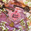 Decorative heart-shaped keychain with 'Fated Mates' text, surrounded by vintage keys and flowers on a textured fabric background.