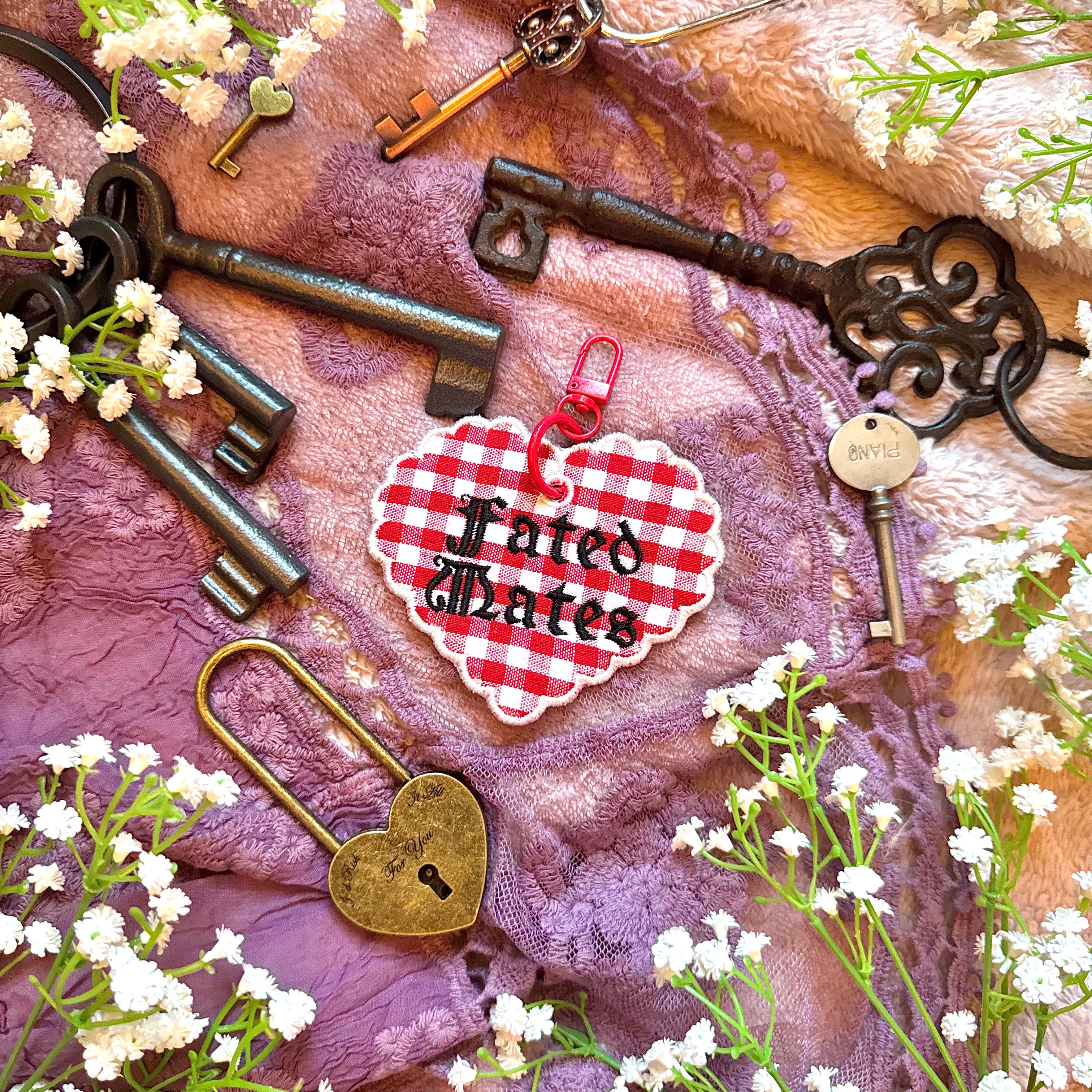 Decorative heart-shaped keychain with 'Fated Mates' text, surrounded by vintage keys and flowers on a textured fabric background.
