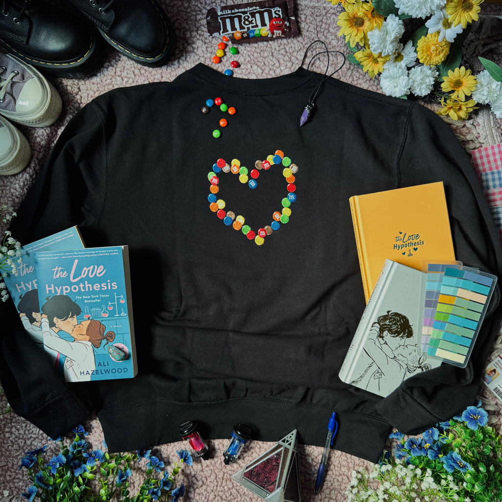 A black crewneck sweatshirt with an embroidered emblem of chocolate candies in the shape of a heart.