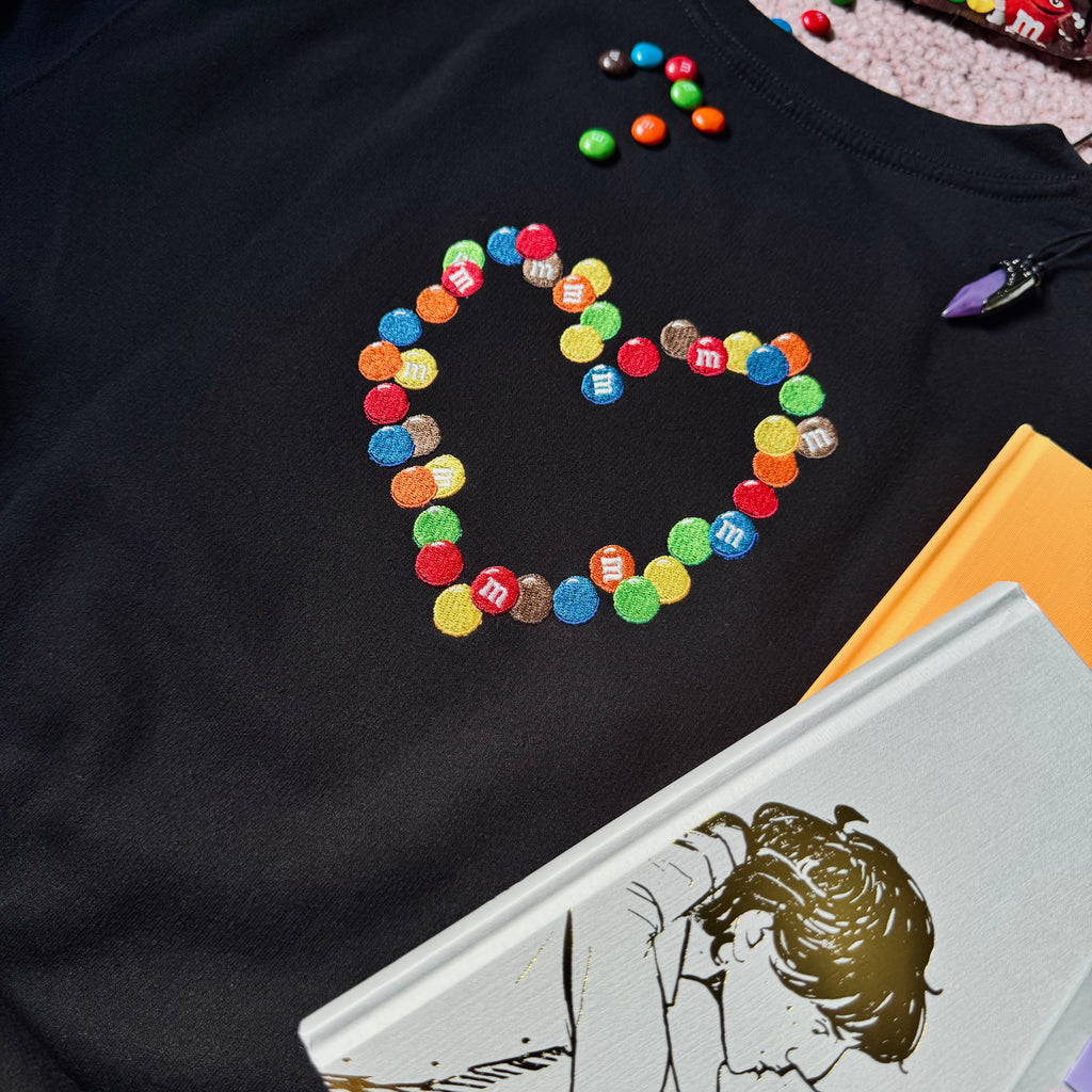 A black crewneck sweatshirt with an embroidered emblem of chocolate candies in the shape of a heart.