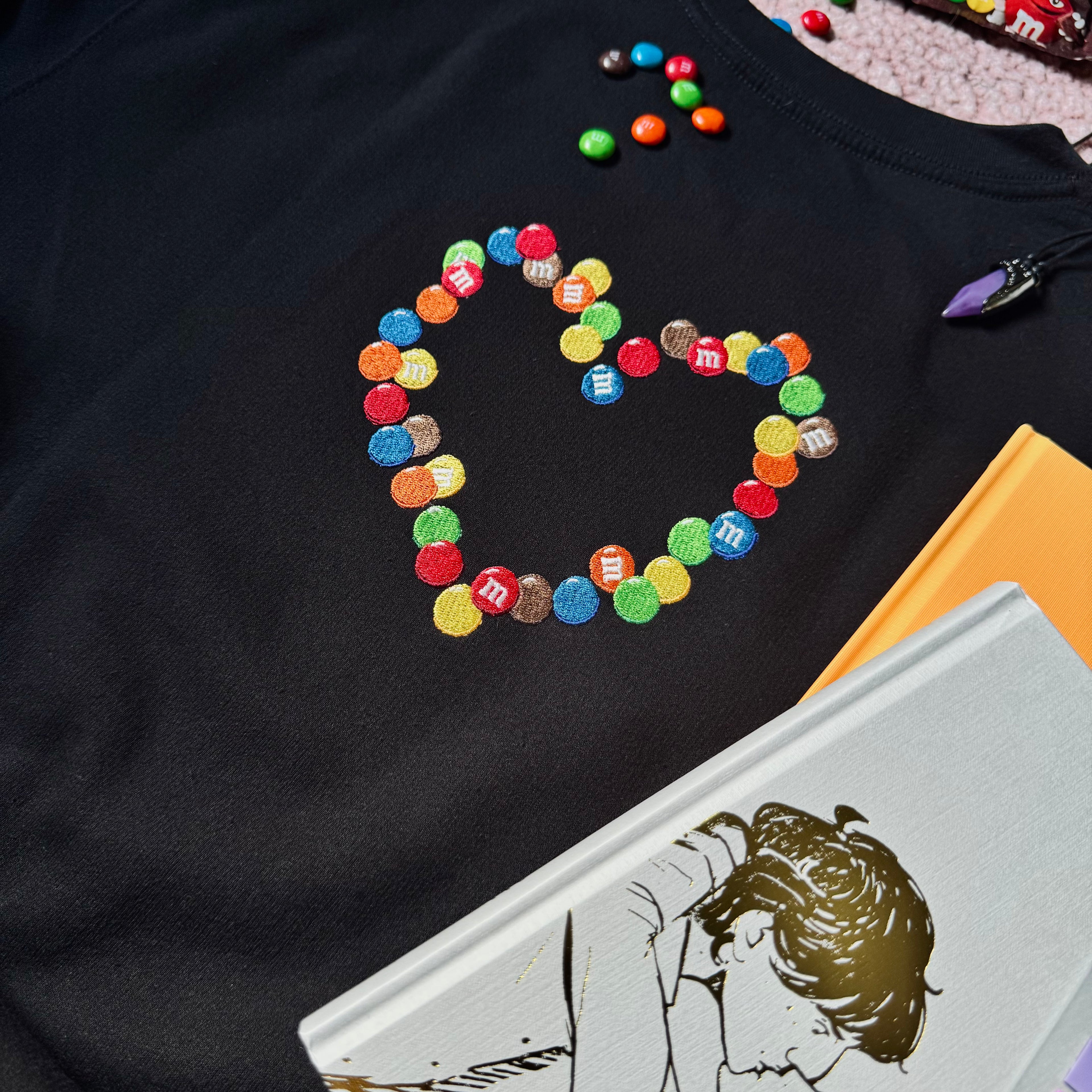 A black crewneck sweatshirt with an embroidered emblem of chocolate candies in the shape of a heart.