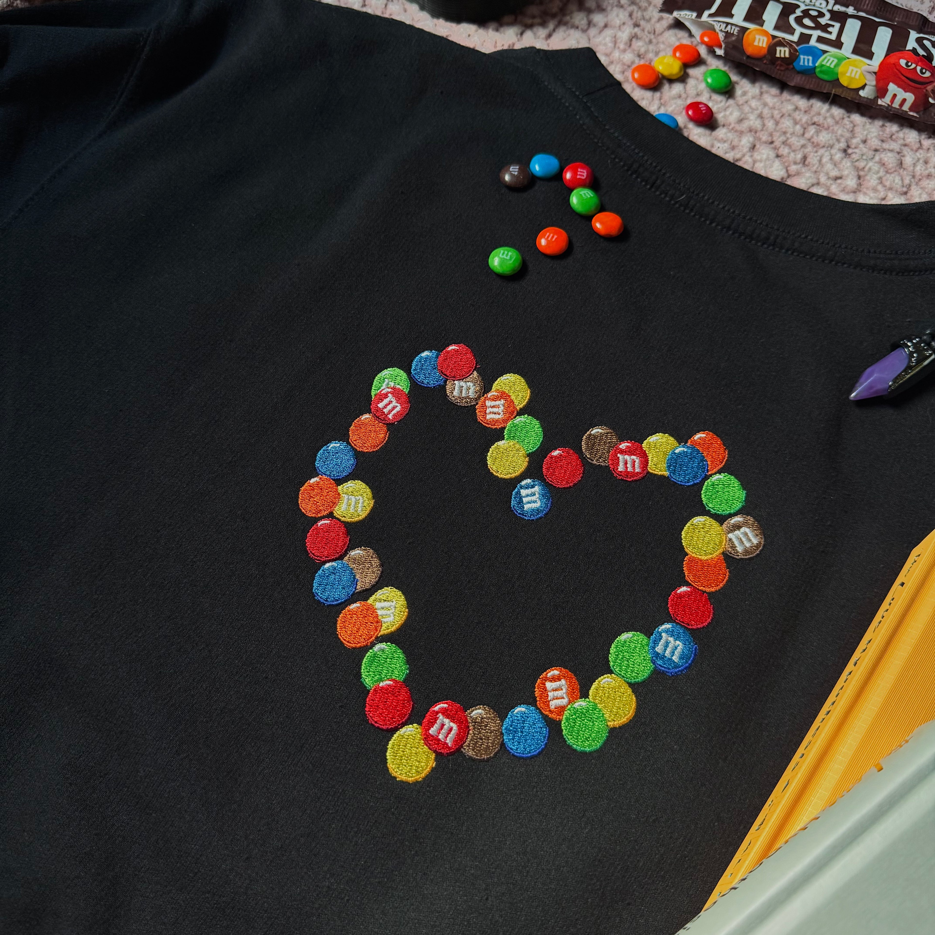 A black crewneck sweatshirt with an embroidered emblem of chocolate candies in the shape of a heart.