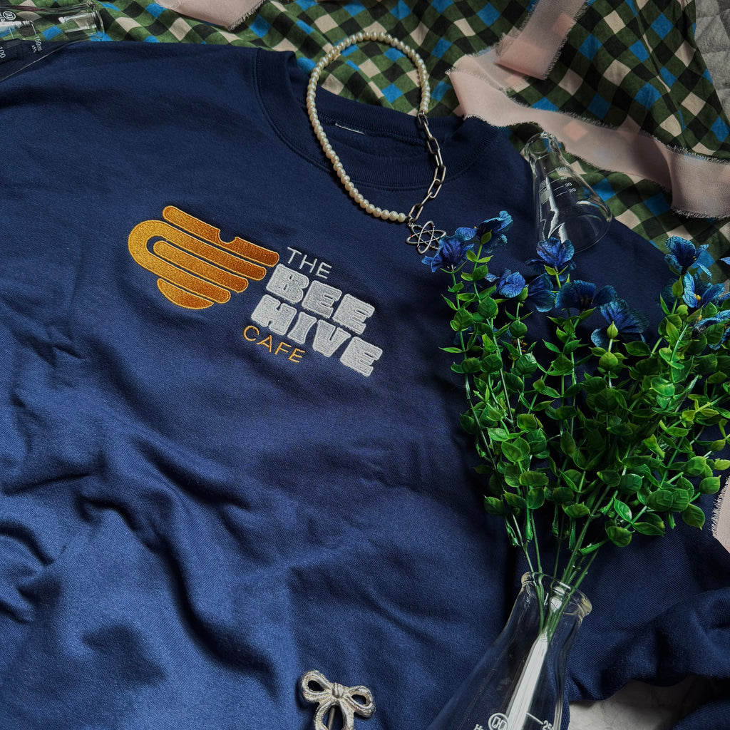 Navy crewneck with embroidered emblem of a mug shaped like an upside down beehive, and the words "The Bee Hive Café."
