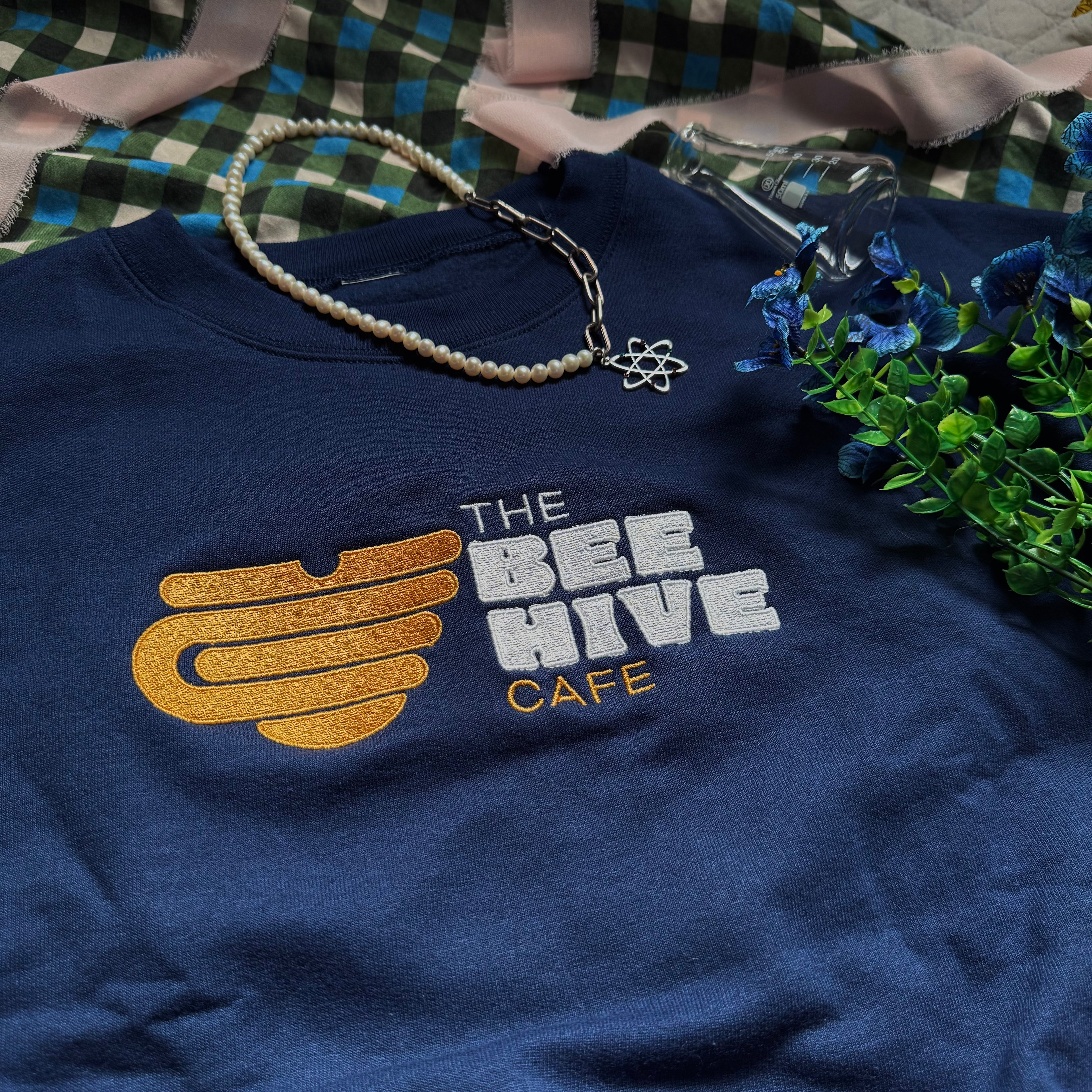 Navy crewneck with embroidered emblem of a mug shaped like an upside down beehive, and the words "The Bee Hive Café."