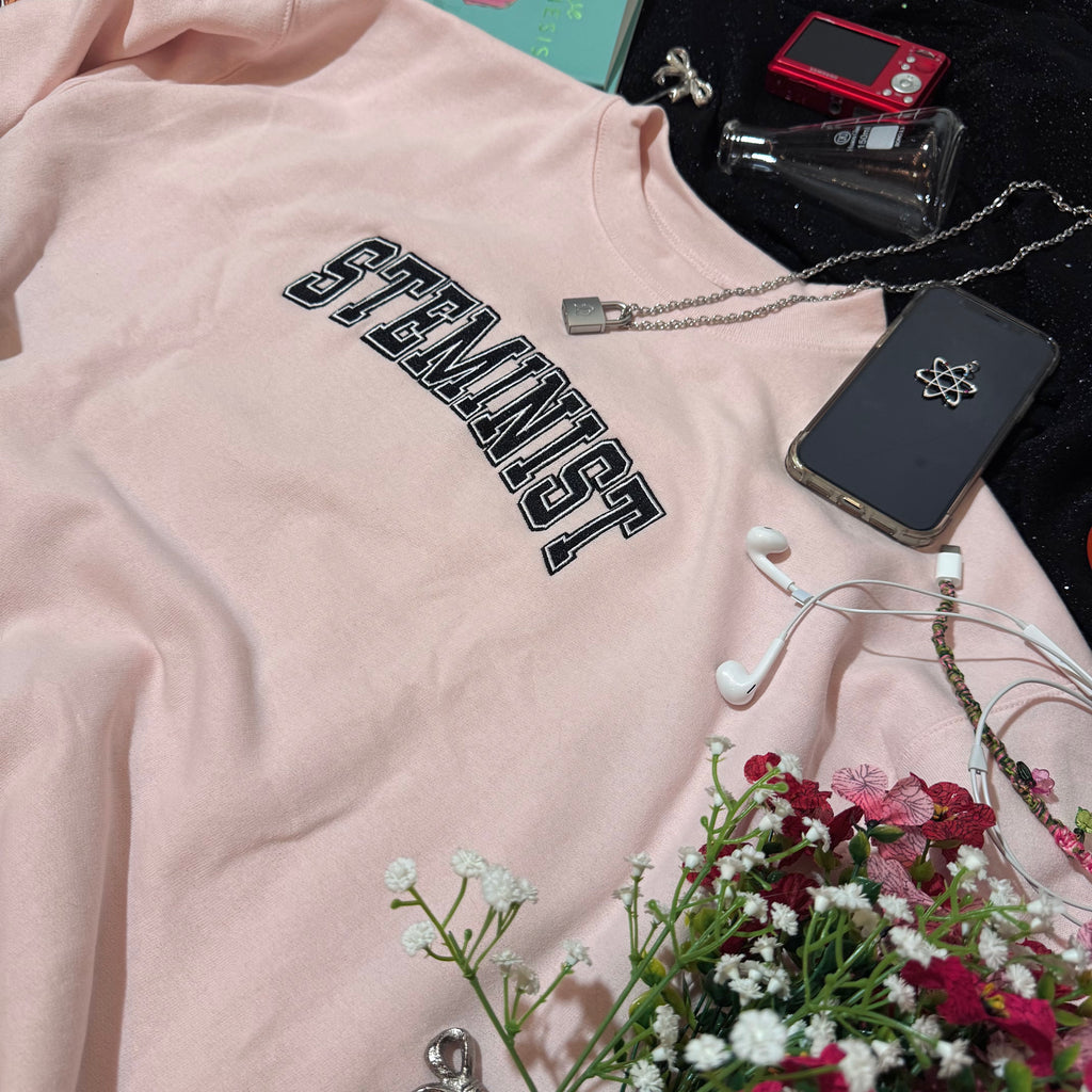 Baby pink sweatshirt with embroidered "STEMINIST" in a varsity font in black and white.