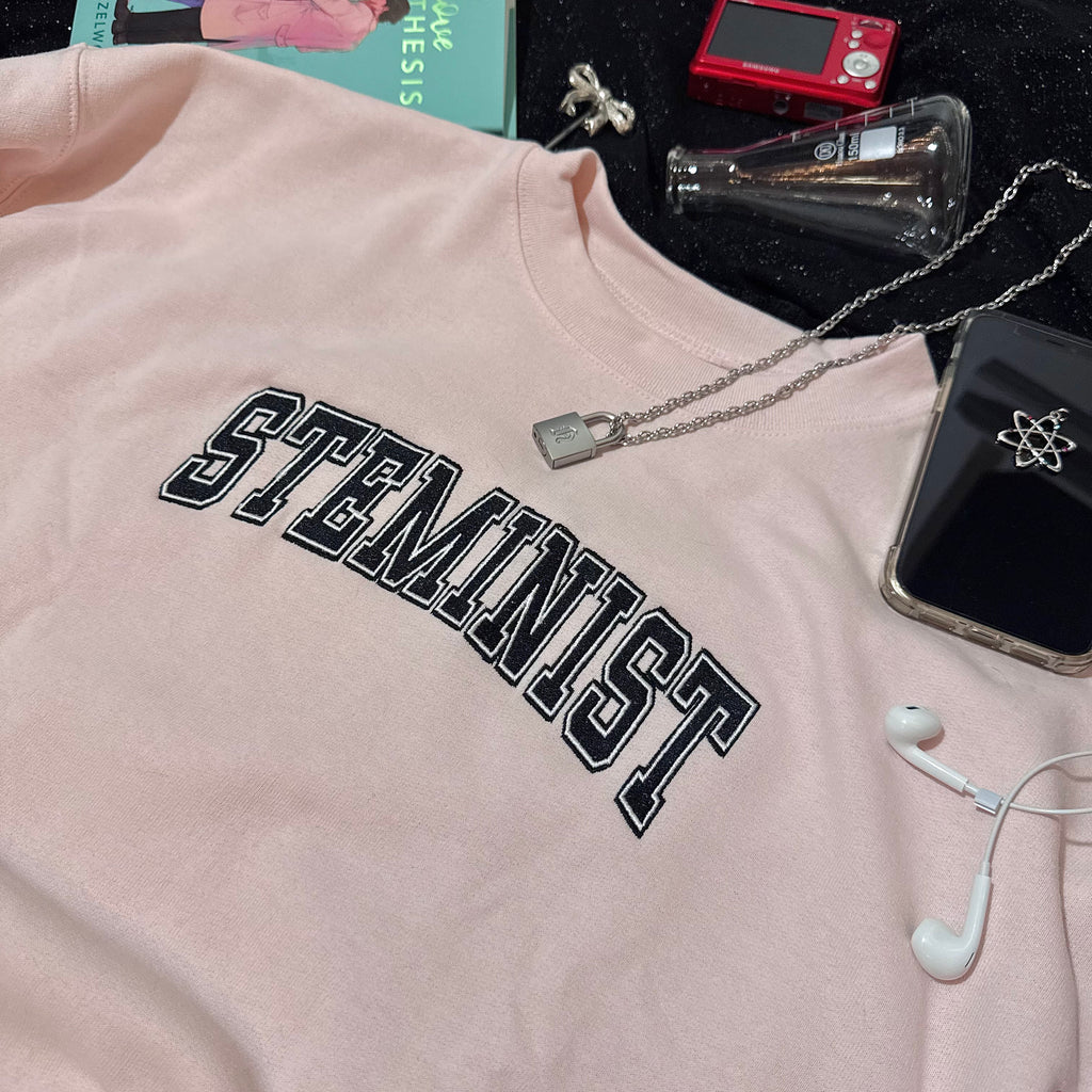 Baby pink sweatshirt with embroidered "STEMINIST" in a varsity font in black and white.